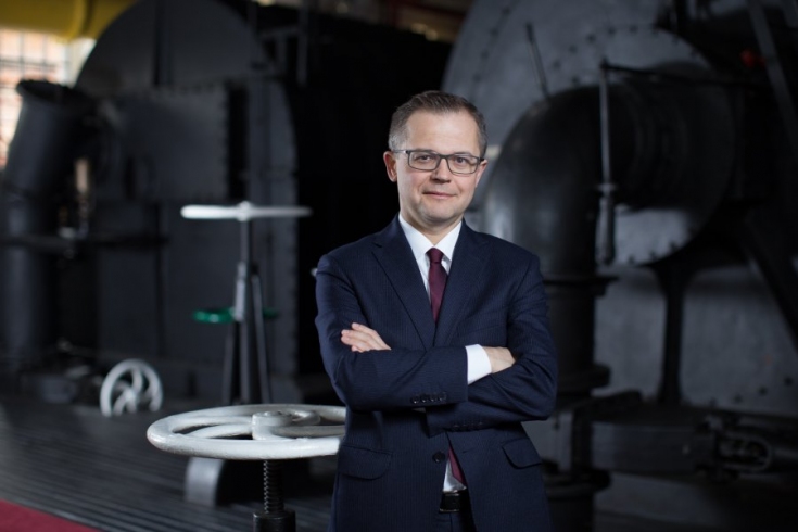 Fot. MSP Absolwent KSAP Wojciech Chmielewski stoi w garniturze z założonymi rękami, w tle środek fabryki